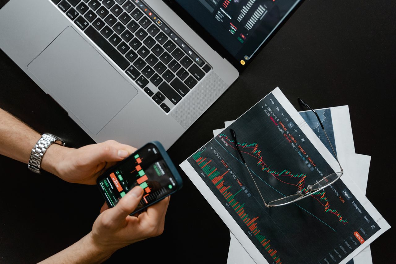 services-01 Hands using smartphone beside laptop with stock charts, showcasing digital trading.